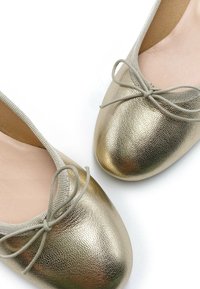 Bailarinas metálicas doradas con superficie texturizada, punta redondeada y un delicado lazo gris que acentúa la parte delantera.
