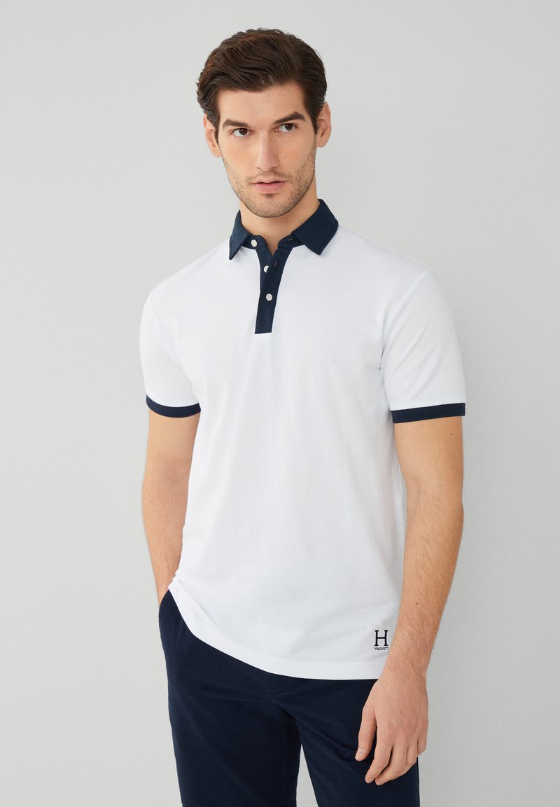 Polo blanc avec un col et des poignets bleu marine, doté d'une patte de boutonnage à trois boutons et d'un détail de logo subtil en bas. Texture de tissu lisse.