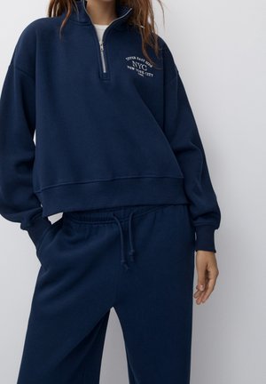 Mujer con sudadera azul marino con media cremallera con el texto "Upper East Side NYC New York City" y pantalones deportivos a juego con cordón.