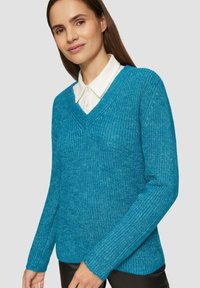 Türkisfarbener Strickpullover mit V-Ausschnitt und geribbelter Textur. Verfügt über lange Ärmel und eine lockere Passform. Darüber getragen über einem weißen Hemd mit Kragen.