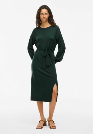 Robe en tricot verte avec une coupe détendue, des manches longues, une ceinture à la taille et une fente sur le côté. Portée avec des sandales à talons marron.
