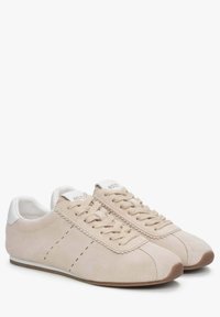 Beżowe zamszowe sneakersy z białymi skórzanymi akcentami, z falistym wzorem w okolicy sznurowania, płaskimi sznurówkami i gumową podeszwą.