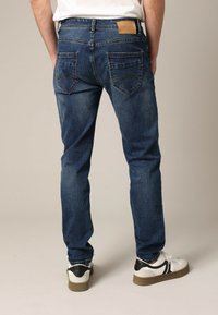 Homme portant un jean bleu foncé et des baskets blanches, se tenant contre un fond beige uni, vu de dos.