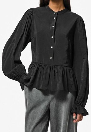Woman wearing black long-sleeve blouse with puffed sleeves, pearl buttons, peplum hem, paired with gray tailored pants.