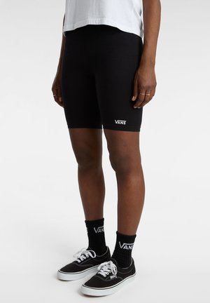 Personne portant un short Vans noir, des chaussettes Vans noires, des baskets Vans noires et blanches, et une chemise blanche, debout devant un fond uni.
