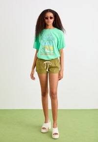 T-shirt verte avec impression graphique de palmiers et de plage, associée à un short olive. Le mannequin porte des sabots à plateforme crème et des lunettes de soleil.