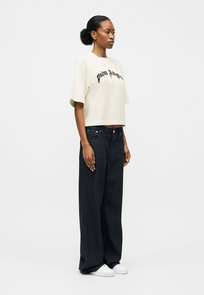 Žena s účesom vo forme drdolu, oblečená v bielom krátkom tričku Palm Angels, širokých čiernych džínsach a bielych teniskách, stojaca zboku.