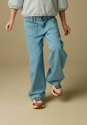 Enfant portant un jean ample bleu clair, un t-shirt rayé, et des baskets roses et beiges, marchant vers l'avant sur un fond beige.