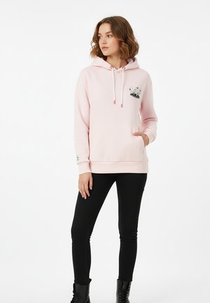 Rosa Kapuzenpullover aus weichem Stoff, mit einer Fronttasche, Kordelzug und einem grafischen Design auf der linken Brust. Kombiniert mit schwarzen Skinny-Jeans.