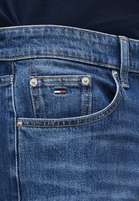 Κοντινή φωτογραφία τσέπης τζιν σε μπλε χρώμα με μικρό ορθογώνιο λογότυπο μάρκας με κόκκινη, λευκή και ναυτικού μπλε ραφή στο ύφασμα.