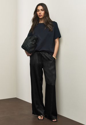 Donna con lunghi capelli castani indossa pantaloni larghi in raso nero, t-shirt oversize blu navy, sandali neri e tiene una grande pochette in pelle nera.