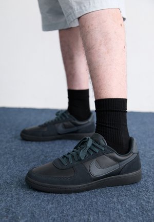 Nogi w jasnych szortach, czarne prążkowane skarpety i czarne buty sportowe Nike stojące na niebieskim wykładzinowym podłodze na tle zwykłej białej ściany.