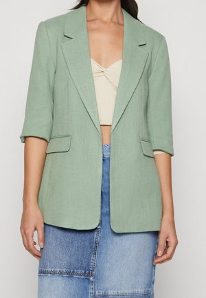 Femme portant un blazer vert clair à manches 3/4, un crop top beige froncé et une jupe patchwork en denim bleu bicolore.