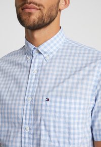 Camicia button-up a quadretti blu chiaro con colletto, dotata di bottoni bianchi e un piccolo logo sulla tasca del petto. Texture liscia.