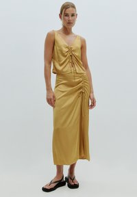 Geel satijnen jurk met een V-hals, geplooide details en een asymmetrische zoom. Gepaard met zwarte flip-flops met een dikke zool.