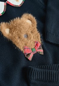 Marineblauwe trui met een tan kleurige, fuzzy teddybeerpatch met crèmekleurige oren en een gemusterd rood en groen strikje, van structuurgebreide stof.