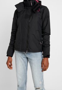 Schwarze Zip-Jacke mit hohem Kragen, Toggle-Details und gerippten Bündchen, mit Seitentaschen und dezentem Markenlogo-Stickerei.