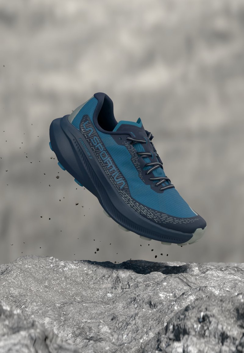 Zapatilla de trail running azul y negra en el aire sobre un terreno rocoso con pequeños escombros flotando alrededor, destacando su uso en exteriores exigentes.