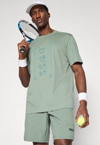 Tricou sportiv de culoare verde deschis, cu logo-ul "BOSS", asociat cu pantaloni scurți asortați. Jucătorul ține o rachetă de tenis și o minge, purtând o brățară albă la încheietură și o șapcă.