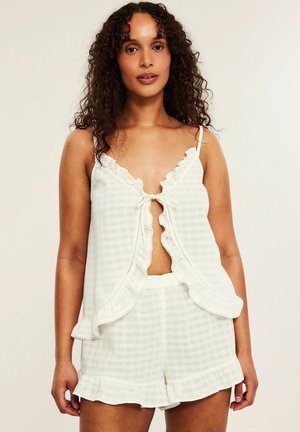 Kvinde med krøllet hår iført hvid ternet flæset ærmeløs top og matchende shorts, stående mod en ensfarvet lys baggrund.