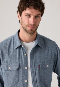Textured blue flannel shirt with two button-flap pockets, white buttons, and a small red logo tab on the left pocket.
