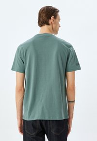 Camiseta de algodón verde menta con mangas cortas, cuello redondo y dobladillo cosido. Presenta un detalle mínimo en la parte posterior. Se lleva con jeans oscuros.