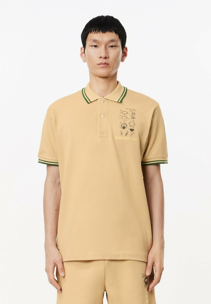 Joven con camisa polo beige con cuello y bordes de mangas a rayas verdes, que presenta un pequeño gráfico y texto en el pecho izquierdo.