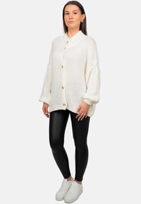 Witte gebreide cardigan met grote knopen, losse fitting en geribbelde manchetten, gecombineerd met zwarte glanzende leggings en witte sneakers.