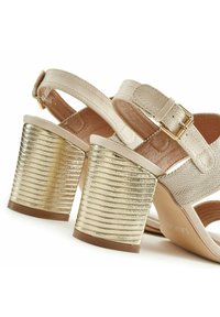 Beige sandalen met visgraatbanden en een dikke, gestructureerde, metallic gouden blokhak. Voorzien van een verstelbare enkelband voor een goede pasvorm.