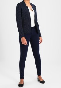 Blazer blu navy con pois bianchi, design aderente, abbinato a una maglietta bianca e jeans skinny blu scuro, completato da ballerine nere.