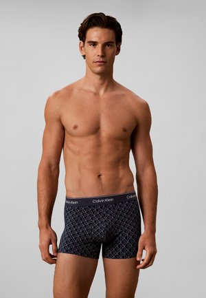 Junger Mann mit kurzen braunen Haaren, der schwarze gemusterte Boxershorts mit Calvin Klein-Logo am Bund trägt, vor einfarbigem grauem Hintergrund.