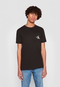Zwart katoenen T-shirt met korte mouwen, ronde halslijn en een linkse borstzak met een wit logo. Casual pasvorm, gladde textuur.