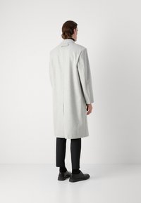 Abrigo de lana gris claro con corte recto y abertura en la espalda, combinado con pantalones y zapatos negros. Diseño simple, con detalles mínimos.