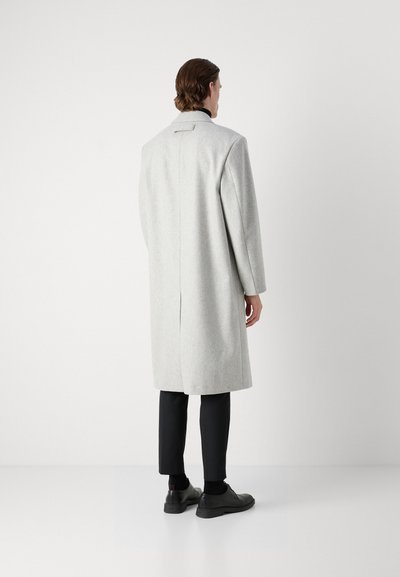 Manteau en laine gris clair avec une coupe droite et une fente au dos, associé à un pantalon et des chaussures noires. Design simple, détails minimaux.