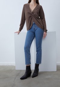 Top marrón de manga larga con detalle de nudo en el frontal, combinado con jeans de mezclilla azules y botas negras hasta el tobillo. El fondo es minimalista y claro.