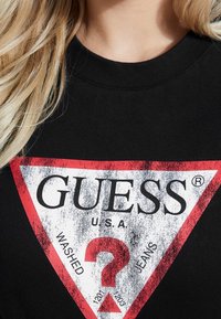 Musta T-paita, jossa on iso, kulunut punavalkoinen kolmiomainen graafinen kuvio, jossa lukee "GUESS," "U.S.A." ja kysymysmerkki.
