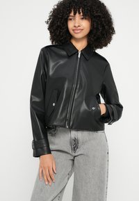 Melnas sintētiskās ādas cropped jaka ar apkakli, rāvējslēdzēja aizdari, divām sānu kabatām un regulējamām aprocēm, kombinēta ar gaiši pelēkiem džinsiem.