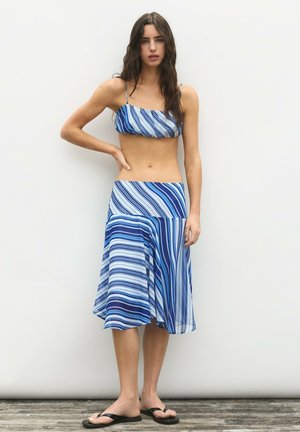 Vrouw met lang donker haar die een blauw-wit gestreepte cropped top en bijpassende midi rok draagt, blootsvoets staand op een houten vloer.