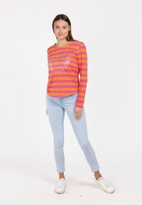 Langärmliges Hemd mit pink-orange gestreiftem Muster und rundem Ausschnitt, kombiniert mit hellblauen Skinny-Jeans und weißen Sneakers.