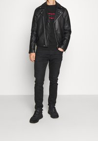 Giacca da motociclista in pelle nera sopra una felpa nera con grafica rossa e bianca, abbinata a jeans scuri e sneakers nere.
