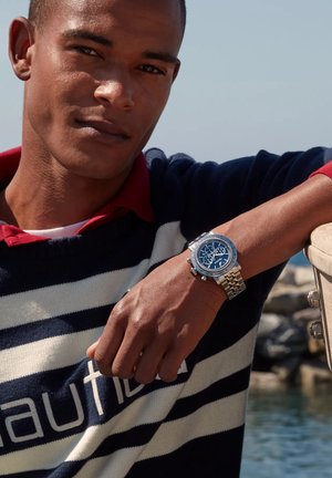Maglione a righe blu e bianche con colletto rosso, indossato sopra una camicia bianca. Orologio argento con quadrante blu e molteplici indicatori sul polso sinistro.