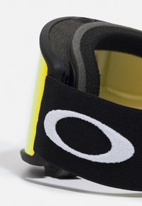 Des lunettes de ski noires avec un revêtement en mousse et une sangle noire ajustable arborant un logo blanc bien visible. La lentille est teintée de jaune avec une finition brillante.