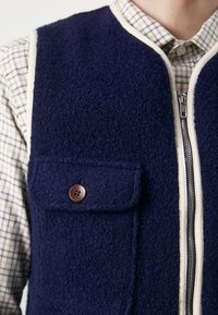 Jūras spēku krāsas flīsa jaka ar krēmīgu apdari, ar priekšpusi ar rāvējslēdzēju, pogu kabatu un strukturētu virsmu. Valkāta virs šūnu krekla.