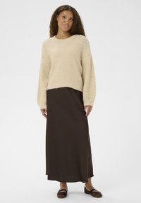 Maglione beige lavorato a maglia con dettagli in perle, maniche larghe e una texture morbida, abbinato a una lunga gonna marrone e scarpe basse marroni.