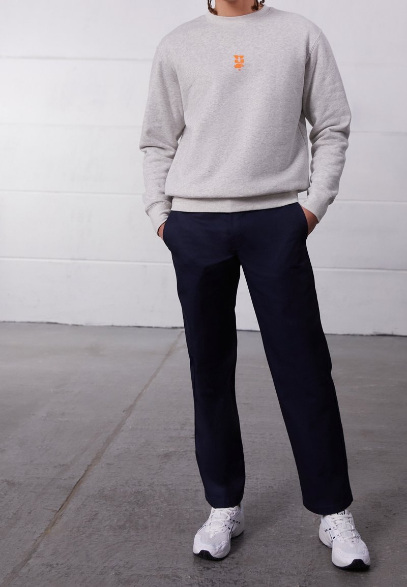 Sweat-shirt gris avec logo orange, associé à un pantalon bleu marine et des chaussures de sport blanches. La tenue présente une coupe décontractée et une texture lisse.