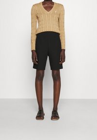 Beige villatakki, jossa on V-pääntie, mustat räätälöidyt shortsit ja mustat liukusandaalit, joissa on geometrinen muotoilu. Sujuva rakenne ja rento tyyli.