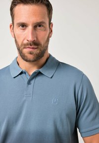 Polo in un delicato colore azzurro, realizzata in tessuto strutturato. Presenta un colletto, chiusura con due bottoni e un logo ricamato sul petto.