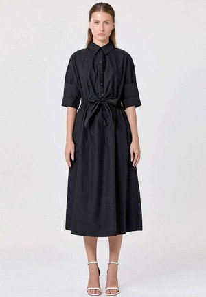Femme portant une robe chemise midi noire avec des manches retroussées et une ceinture nouée à la taille, associée à des talons blancs à bride cheville, debout sur un fond uni.