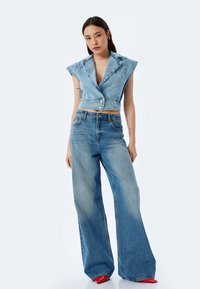 Gilet di jeans tagliato con ampie rever e bottoni frontali, abbinato a jeans azzurri a gamba larga. Tacchi rossi a punta e una texture liscia.