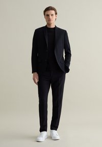 Completo nero su misura con un design elegante, abbinato a una maglietta nera e sneakers bianche. Tessuto morbido, taglio dritto e dettagli minimi.